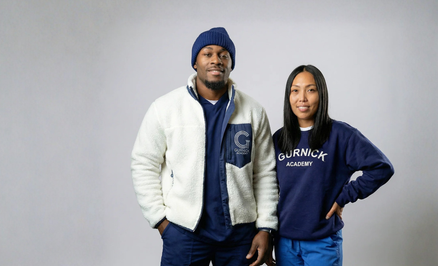 Two people pose together in blue and navy outfits with Gurnick Academy logos visible on their jackets and shirts against a gray backdrop.